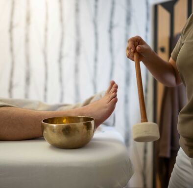Mann liegt auf Liege, Masseurin schlägt eine kleine Klangschale, die neben ihm steht, mit einem Schläger an.
