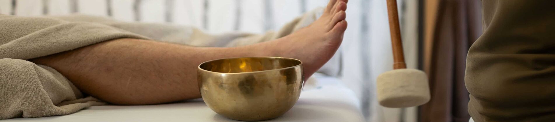 Mann liegt auf Liege, Masseurin schlägt eine kleine Klangschale, die neben ihm steht, mit einem Schläger an.