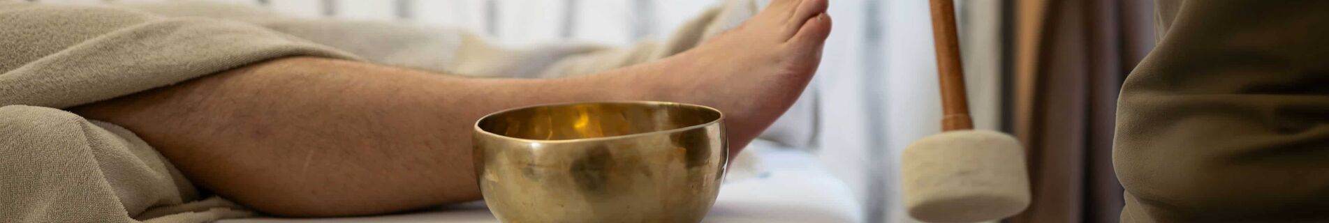 Mann liegt auf Liege, Masseurin schlägt eine kleine Klangschale, die neben ihm steht, mit einem Schläger an.