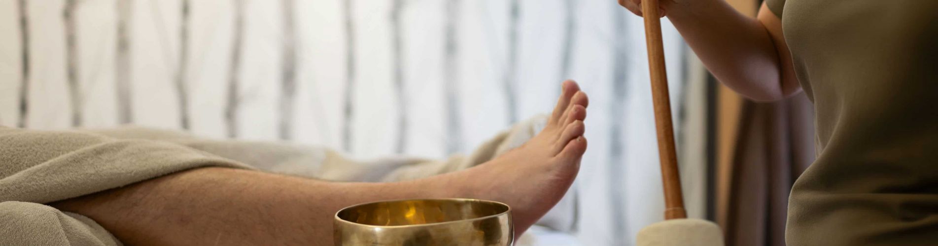 Mann liegt auf Liege, Masseurin schlägt eine kleine Klangschale, die neben ihm steht, mit einem Schläger an.